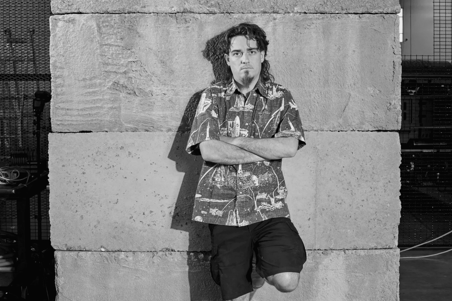 A person with curly hair and a goatee is leaning against a large stone wall. They are wearing a patterned shirt and shorts, with arms crossed, looking directly at the camera. The setting appears to be industrial or urban. The image is in black and white.